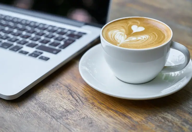 A cup of cappuccino next to a laptop on a wood grain desk